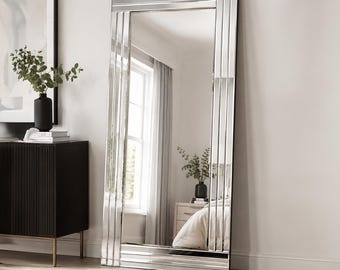 Op de afbeelding: Een hoge, rechthoekige vloerspiegel met een brede, uit meerdere panelen bestaande lijst met een reflecterende spiegelafwerking. De lijst bestaat uit diverse verticale en horizontale glasstroken die een gelaagde, geometrische rand vormen.