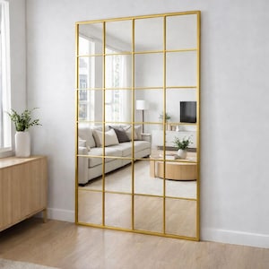 May include: A large rectangular floor mirror featuring a thin gold metal frame divided into a grid of twenty square panes. The mirror reflects a living room interior with a sofa, a coffee table, and a television set, creating a window-like effect.