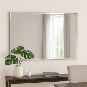 May include: A rectangular wall mirror with a thin, metallic frame. The mirror reflects a window with white curtains. Below the mirror, a dark brown console table holds a white vase with a green monstera plant, a small white cup, and two books.