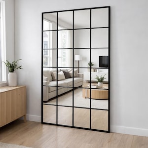 May include: A large rectangular floor mirror featuring a black metal grid frame that divides the glass into sixteen equal square panes. The mirror reflects a living room interior, showing a sofa, a coffee table, and a television stand.