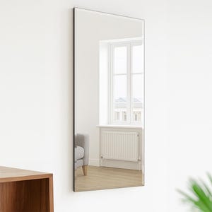 May include: A long, rectangular mirror with no frame, mounted on a white wall. The mirror reflects a room with a window, radiator, and a grey sofa. The mirror is approximately 152 cm high and 51 cm wide.
