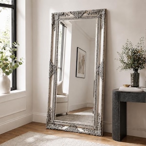 May include: A large rectangular floor mirror featuring an ornate, cream-coloured frame with intricate, dark-toned decorative carvings at the corners and along the edges. The mirror has a reflective glass surface that shows a room interior.