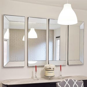 May include: Four rectangular wall mirrors with bevelled edges arranged in a row. Each mirror features a thin black frame. The mirrors reflect a room with a white pendant light, a patterned wall, and a doorway. The mirrors are mounted on a wall.