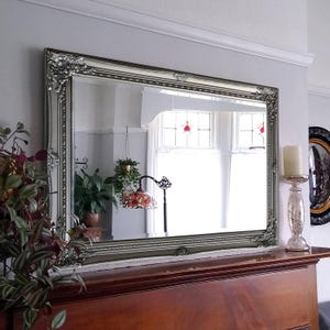 May include: A rectangular wall mirror featuring an ornate, silver-toned frame with intricate scrollwork and decorative corner detailing. The mirror is mounted above a wooden mantelpiece, reflecting a room interior with a lamp and plants.