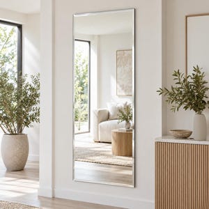 May include: A tall, rectangular wall mirror featuring a slim, polished silver metal frame. The mirror is mounted vertically on a plain white wall, reflecting a bright living space that includes a beige sofa, a wooden side table, and a plant.