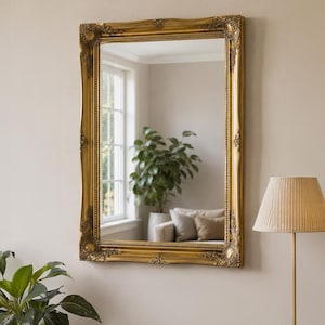 May include: A rectangular wall mirror featuring an ornate gold frame with intricate carved detailing at the corners and along the edges. The reflective glass surface displays a clear view of an interior room with a window, a plant, and furniture.