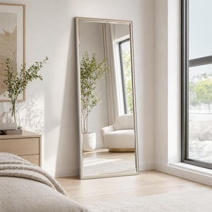 May include: A tall, rectangular floor mirror with a slim, metallic silver frame. The mirror is positioned upright against a wall, reflecting a bright interior space that includes a potted green plant and a cream-coloured armchair.