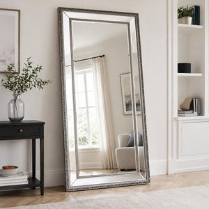 May include: A tall, rectangular floor mirror featuring a wide, ornate silver frame with intricate detailing. The mirror is positioned leaning against a wall, reflecting a bright room with a window, white curtains, and a light-coloured upholstered chair.