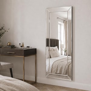 May include: A tall, rectangular wall mirror featuring a wide, bevelled glass frame with a reflective silver finish. The mirror is mounted vertically on a plain wall, displaying a clear reflection of a bedroom interior with a bed and window.