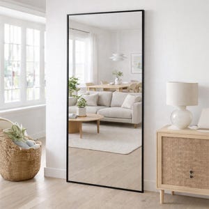 May include: A tall, rectangular floor mirror featuring a thin, black metal frame. The mirror is positioned against a white wall, reflecting a bright living room space that includes a light-coloured sofa, a wooden coffee table, and a dining area.