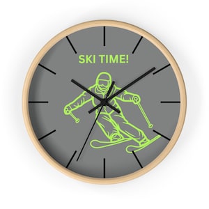 May include: A gray wall clock with a wooden frame features a green line drawing of a skier on a slope. The clock face has black hour and minute hands and black markings for the hours. The text "SKI TIME!" is printed in green on the clock face.