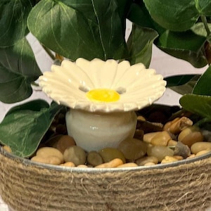 Puede incluir: Una maceta decorativa con una parte superior de cerámica en forma de margarita y un centro amarillo. La maceta está llena de piedras pequeñas y rodeada de hojas verdes. La maceta está envuelta con cuerda.