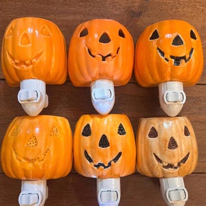 May include: Six ceramic pumpkin nightlights with different facial expressions. The pumpkins are orange and have black facial features. The nightlights have a white base with a switch.