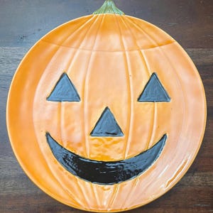 以下が含まれることがあります： オレンジ色のセラミック製の、かぼちゃの形をしたサービングプレート。黒く塗られたジャック・オ・ランタンの顔が描かれている。プレートは光沢のある仕上げになっている。