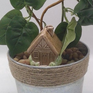 May include: A miniature ceramic house with a brown thatched roof and a chimney sits in a galvanized metal pot. The pot is wrapped with twine and filled with small stones. Green leaves and vines surround the house, creating a miniature garden scene.