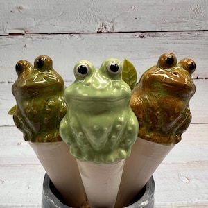 Może przedstawiać: Trzy ceramiczne figurki żab z zieloną i brązową glazurą. Żaby siedzą na białych ceramicznych stożkach. Stożki znajdują się w szarym garnku.