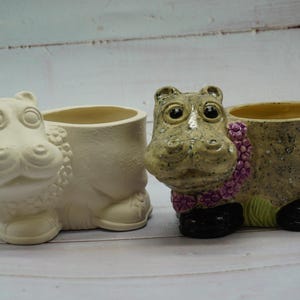 May include: Two ceramic hippopotamus planter pots. One is white and the other is beige with brown speckles and a purple flower garland. Both have black shoes.