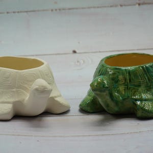 Puede incluir: Dos macetas de cerámica en forma de tortuga, una blanca y otra verde con un caparazón estampado. Ambas macetas tienen una abertura redonda para plantar.