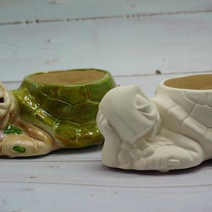May include: Two ceramic turtle planters, one green and one white, with a flower design on the head. Both are lying on their backs with their heads turned to the side.