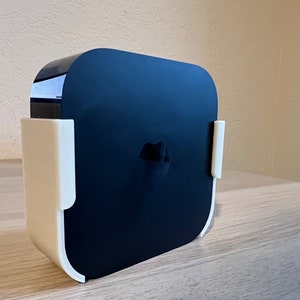 Op de afbeelding: Een zwart Apple TV streamingapparaat met een wit Apple-logo aan de voorkant, gemonteerd op een witte wandbeugel.