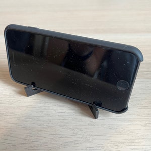 May include: A black smartphone is propped up on a black plastic stand. The phone is in a black case and has a black screen.