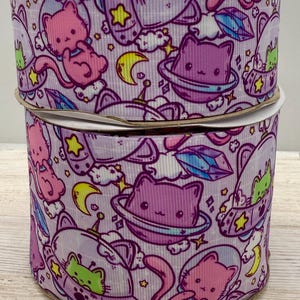 May include: Two rolls of purple and white patterned ribbon with a space cat theme. The ribbon features cats in spaceships, planets, stars, and moons.