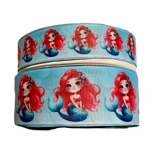 Può includere: Un nastro blu con un motivo ripetuto di sirene dai capelli rossi e code blu. Le sirene sorridono e hanno dei fiori nei capelli.