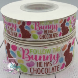 Könnte beinhalten: Zwei Spulen weißes Band mit Ostermotiv. Das Band zeigt ein sich wiederholendes Muster aus braunen Hasen, bunten Tupfen und dem Text "Follow the Bunny, He Has Chocolate" in Pink und Grün.