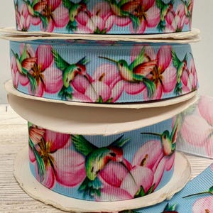 May include: Spools of decorative ribbon with a repeating pattern of hummingbirds and pink flowers on a light blue background. The ribbon is stacked, with a piece unrolled at the bottom. Ideal for crafts.