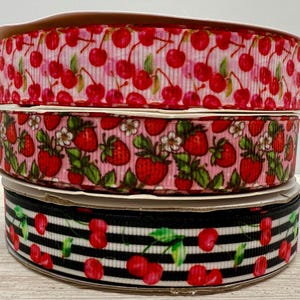 Könnte beinhalten: Drei Rollen dekoratives Band. Die obere Rolle hat einen rosa Hintergrund mit einem Kirschmuster. Die mittlere Rolle hat einen rosa Hintergrund mit Erdbeeren. Die untere Rolle hat einen schwarz-weiß gestreiften Hintergrund mit Kirschen.