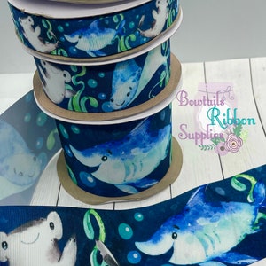 May include: A stack of blue and white ribbon with a watercolour design of sharks and stingrays. The ribbon is on a white surface with the text "Bowtails Ribbon Supplies" in the background.