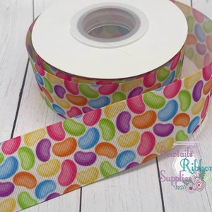 May include: A spool of white ribbon with a jelly bean pattern in various colours, including pink, purple, blue, green, orange, and yellow. The ribbon is on a white wooden surface.