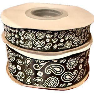 Könnte beinhalten: Zwei Spulen schwarzes Band mit weißem Paisley-Muster. Das Band ist gestapelt und zeigt das detaillierte Design. Die Spulen haben einen neutralfarbenen Kern und Kanten, die einen Kontrast zum kräftigen Muster bilden. Ideal für Bastel- und Dekorationsprojekte.