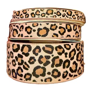 May include: Several rolls of ribbon with a leopard print design. The ribbon has a beige base with brown and black spots. The rolls are different widths, with the widest at the bottom and the narrowest at the top.