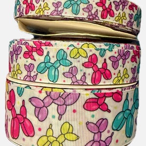 Könnte beinhalten: Ein Stapel aus drei Rollen weißem Ripsband mit einem sich wiederholenden Muster aus Luftballontieren in den Farben Pink, Gelb, Lila und Türkis, akzentuiert durch kleine bunte Punkte, die über die Stoffoberfläche verteilt sind.