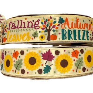 Könnte beinhalten: Zwei Spulen dekorativen Bandes. Die obere Spule hat die Worte "falling leaves" und "Autumn Breeze" mit einer Baum- und Kürbisgrafik. Die untere Spule hat ein Muster aus Sonnenblumen, Blättern und Eicheln.