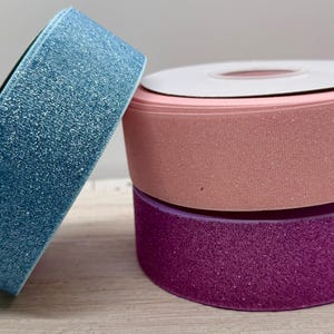 May include: Three rolls of glitter ribbon in shades of blue, pink, and purple. The blue ribbon leans against the other two rolls. The pink ribbon has a white cardboard centre. The ribbons are on a light wood surface.