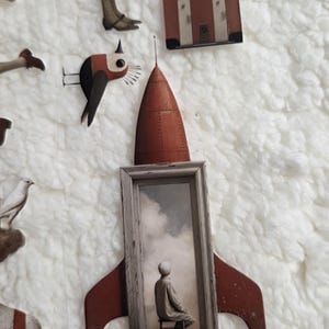 May include: A collage of paper cutouts featuring a red rocket with a framed image of a person, a bird, a suitcase, and a figure in a dress. The background is a fluffy white texture.