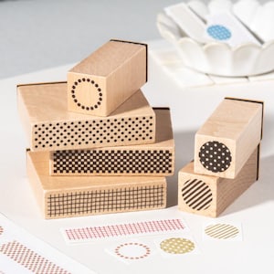 Peut inclure: Une collection de tampons en bois présentant divers motifs géométriques, notamment des points, des cercles, des grilles et des rayures. Les tampons sont des blocs rectangulaires et carrés avec des motifs à l'encre noire. Des échantillons de papier sont disposés devant.