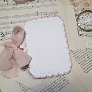 Könnte beinhalten: Eine leere Karte mit einem rosa, wellenförmigen Rand und einer rosa Schleife. Zwei Rollen rosa Washi-Tape und eine Vintage-Kamera sind ebenfalls zu sehen. Trockenblumen und Notenblätter vervollständigen die Ästhetik.