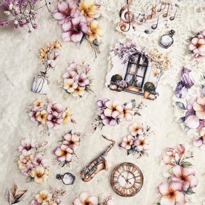 Può includere: Un assortimento di adesivi floreali ed elementi decorativi, tra cui fiori nei toni del rosa, giallo e viola, un sassofono, un orologio e note musicali. Gli adesivi sono disposti su una superficie strutturata bianco sporco.