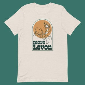 May include: A beige t-shirt with a graphic of a person playing drums in a green and orange arch. The text "more Levon" is below the graphic.