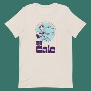 Puede incluir: Una camiseta blanca con una ilustración de estilo vintage de J.J. Cale tocando la guitarra frente a un micrófono. El texto "J.J. Cale" está en una fuente retro en negrita.
