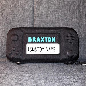 May include: Black carrying case for a video game console with a blue and white label that says "BRAXTON CUSTOM NAME".