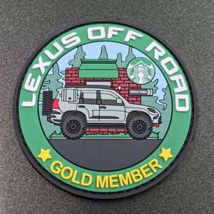 May include: A circular rubber patch featuring a light gray SUV parked in front of a brick building with a green awning and a circular logo. The text reads LEXUS OFF ROAD and GOLD MEMBER, with two yellow stars flanking the bottom text.
