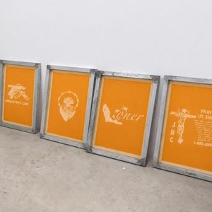 May include: Four rectangular screen printing frames with weathered grey wooden borders and vibrant orange mesh. The frames display white graphic prints including a handgun with the text HANDLE WITH CARE, a sacred heart, the word loner, and a crucifix.