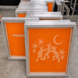 May include: A stack of rectangular aluminium screen printing frames with bright orange mesh. The front frame features a light-coloured graphic of three stylised figures dancing under a crescent moon and stars, while another frame shows the text PDB.