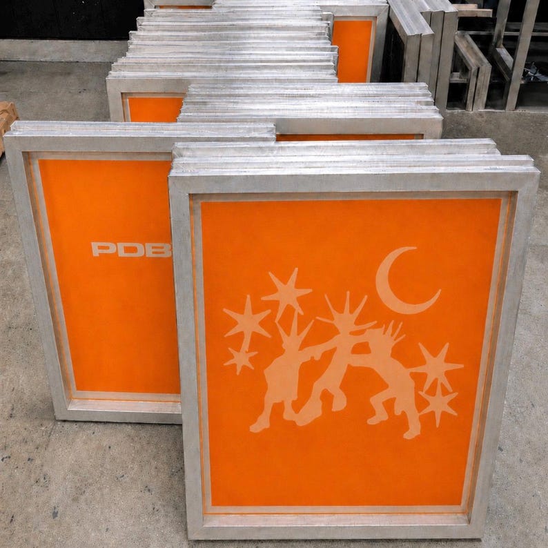 May include: A stack of rectangular aluminum screen printing frames with bright orange mesh. The front frame features a light-colored graphic of three stylized figures dancing under a crescent moon and stars, while another frame shows the text PDB.