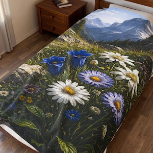 May include: A blanket with a floral design, depicting a mountain scene and a meadow of wildflowers. The blanket features large white daisies, bluebell-like flowers, and purple daisies with yellow centres. The background shows a mountain range under a blue sky.