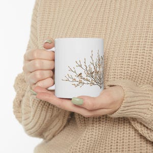 May include: White ceramic mug featuring a nature-inspired design. The mug showcases a detailed illustration of a tree branch with small birds perched on it. The artwork is in shades of brown and beige, creating a natural aesthetic. The mug has a standard handle.